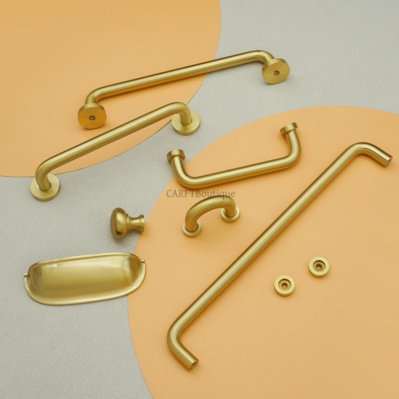 May include: A selection of brass-toned cabinet hardware, including handles, knobs, and a small tray. The hardware has a smooth, matte finish and a simple, modern design. The pieces are arranged on a peach and grey background.