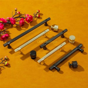 May include: A collection of decorative cabinet hardware, including black and silver drawer pulls and knobs. The pulls have a textured diamond pattern, and the knobs have a similar design. The hardware is displayed on a mustard-colored surface, with a floral arrangement in the background.