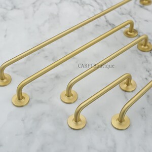 May include: A collection of gold-colored cabinet pulls in various sizes. Each pull features a curved handle and circular mounting plates. The pulls are arranged on a marble-patterned surface. The text "CARFTBoutique" is visible.