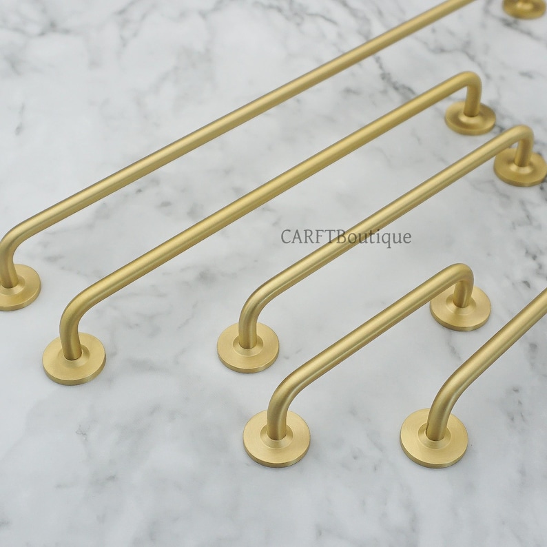 May include: A collection of gold-colored cabinet pulls in various sizes. Each pull features a curved handle and circular mounting plates. The pulls are arranged on a marble-patterned surface. The text "CARFTBoutique" is visible.