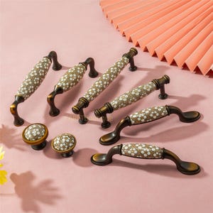 以下が含まれることがあります： キャビネット用の金具のコレクション。取っ手とノブが含まれています。取っ手はブロンズ仕上げで、クリーム色の花柄のセラミックセンターが付いています。ノブも同様のデザインです。ピンク色の表面に金具が展示されています。