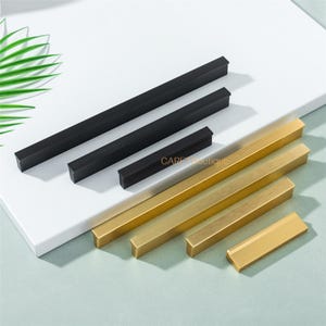 Może przedstawiać: Zestaw prostokątnych uchwytów do szafek w kolorze czarnym i złotym. Uchwyty różnią się rozmiarem i są ułożone na białej powierzchni. Widoczny jest napis "CARPE boutique". Uchwyty są wykonane z metalu.