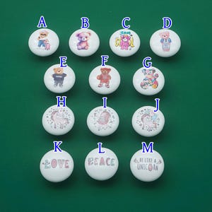 May include: A collection of white ceramic knobs with various designs. The knobs feature illustrations of teddy bears, unicorns, and inspirational words like "LOVE", "PEACE", and "BE LIKE A UNICORN". The knobs are set against a green background.