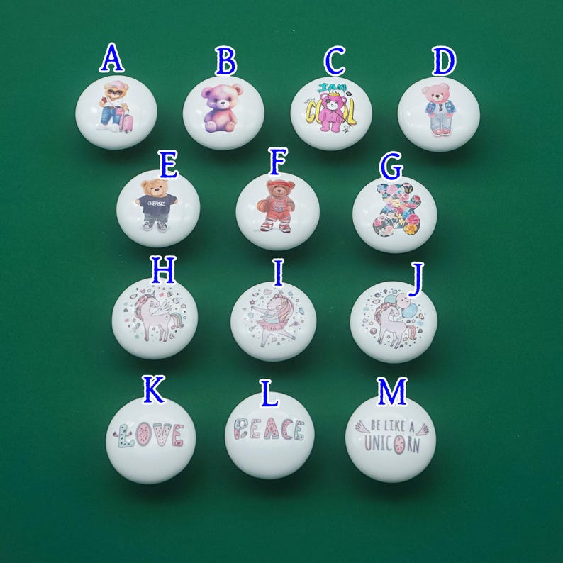 May include: A collection of white ceramic knobs with various designs. The knobs feature illustrations of teddy bears, unicorns, and inspirational words like "LOVE", "PEACE", and "BE LIKE A UNICORN". The knobs are set against a green background.