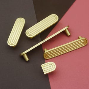 Puede incluir: Juego de herrajes dorados para armarios. El juego incluye manijas y pomos de varios tama&ntilde;os con un dise&ntilde;o ovalado acanalado. El hardware se muestra sobre un fondo de dos tonos marr&oacute;n y rosa.