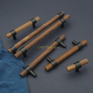 以下が含まれることがあります： 黒い金属アクセントが付いた木製キャビネットプル。プルはさまざまな長さで、円筒形の木製ハンドル、黒い金属リング、T字型のベースが特徴です。「CARFTBoutique」の文字が見えます。