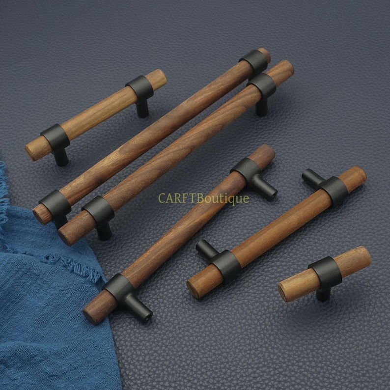 以下が含まれることがあります： 黒い金属アクセントが付いた木製キャビネットプル。プルはさまざまな長さで、円筒形の木製ハンドル、黒い金属リング、T字型のベースが特徴です。「CARFTBoutique」の文字が見えます。