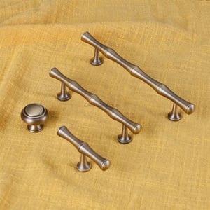 K&ouml;nnte beinhalten: Ein Satz antiker bronzefarbener Schrankbeschl&auml;ge, darunter zwei lange Griffe, ein kurzer Griff und ein runder Knopf. Die Beschl&auml;ge haben ein bambusartiges Design und sind vor einem gelben Stoffhintergrund platziert.