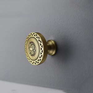 May include: A brass cabinet knob with a circular design. The knob features a central floral pattern surrounded by concentric rings of geometric shapes. The knob is mounted on a gray surface, showcasing its decorative details.