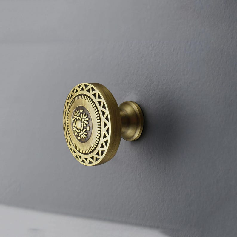 May include: A brass cabinet knob with a circular design. The knob features a central floral pattern surrounded by concentric rings of geometric shapes. The knob is mounted on a gray surface, showcasing its decorative details.