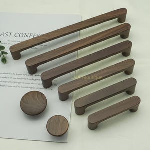 以下が含まれることがあります： さまざまなサイズの木製キャビネットプルとノブのコレクション。プルは丸みを帯びた端を持つ長方形で、ノブは円形です。木材は温かみのある茶色です。アイテムは白い表面に配置されています。