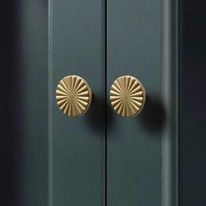 May include: Two gold-coloured cabinet knobs with a sunburst design are mounted on a dark teal cabinet door. The knobs are circular with radiating lines, creating a textured, decorative element. The cabinet doors are vertical and have a smooth finish.