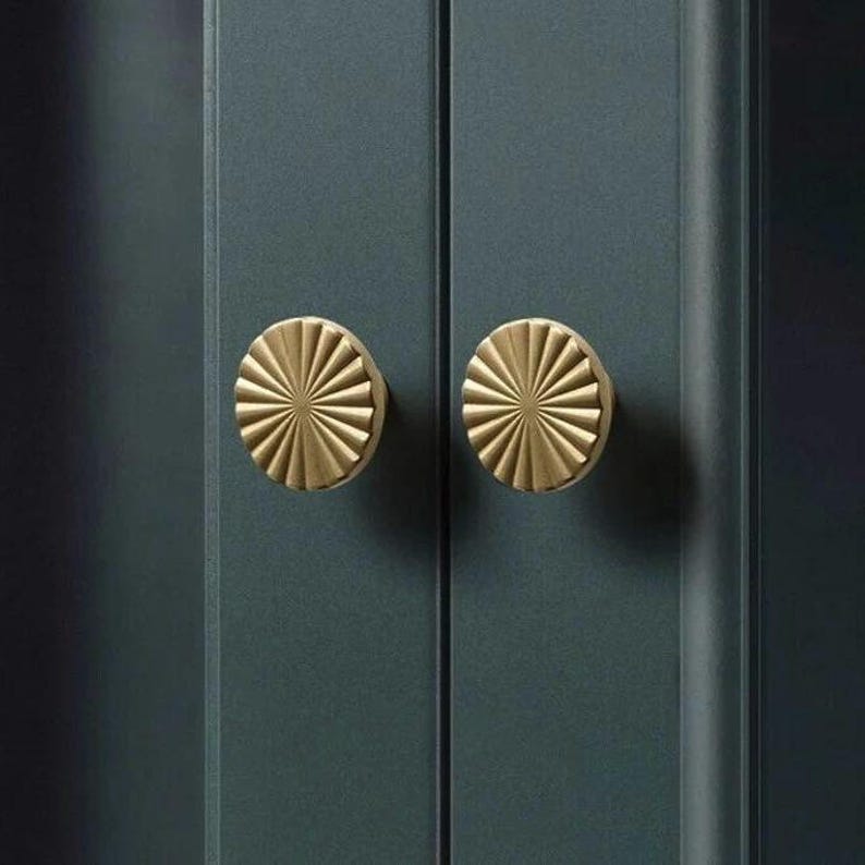 May include: Two gold-coloured cabinet knobs with a sunburst design are mounted on a dark teal cabinet door. The knobs are circular with radiating lines, creating a textured, decorative element. The cabinet doors are vertical and have a smooth finish.