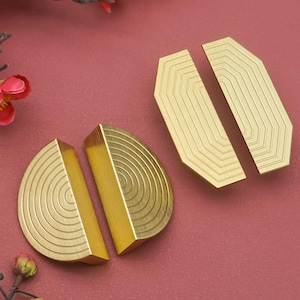 May include: Set of four brass cabinet pulls. Two are semi-circular with concentric grooves, and two are rectangular with parallel lines. The pulls have a gold finish and are displayed on a red background. The pulls are likely for furniture.