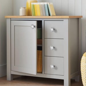 May include: A grey cabinet with a light wood top and silver starburst knobs. The cabinet has a door and three drawers. The door is open, revealing the interior. Books and decorative items are on top.