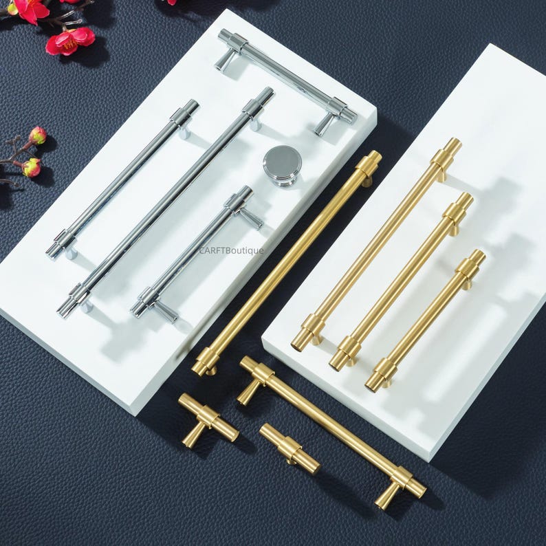 Peut inclure: Divers assortiments de quincaillerie d'armoires, comprenant des poign&eacute;es et des boutons argent&eacute;s et dor&eacute;s. Les poign&eacute;es sont de style barre et en forme de T, de diff&eacute;rentes longueurs. Le mat&eacute;riel est pr&eacute;sent&eacute; sur des plateformes blanches.