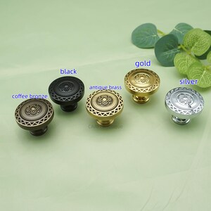 May include: A collection of decorative cabinet knobs in various finishes: coffee bronze, black, antique brass, gold, and silver. Each knob features a circular design with a central floral pattern and a geometric border. The knobs are displayed on a light green surface.