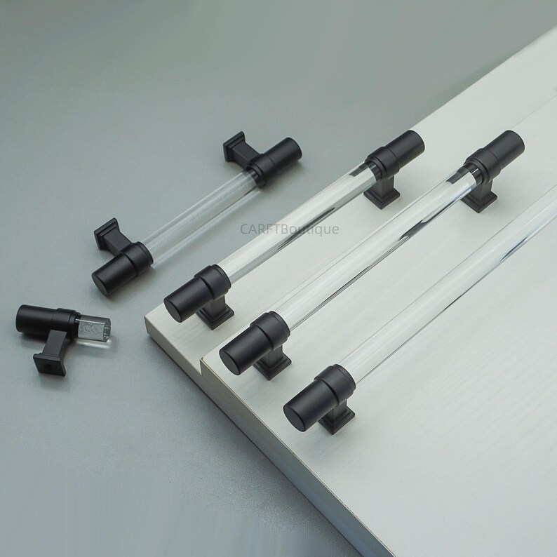 K&ouml;nnte beinhalten: Schrankgriffe aus klarem Acryl und schwarzem Metall. Die Griffe haben einen zylindrischen Acrylmittelteil und schwarze Metallenden und -sockel. Der Text "CARTBOque" ist auf dem Bild sichtbar. Diese Griffe eignen sich f&uuml;r K&uuml;chen- oder Badezimmerschr&auml;nke.