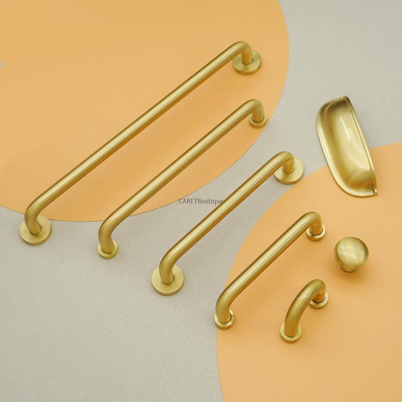 May include: A selection of gold-coloured cabinet hardware, including bar pulls of different lengths, a cup pull, a knob, and a small curved pull. The hardware is arranged on a two-tone beige and orange background.