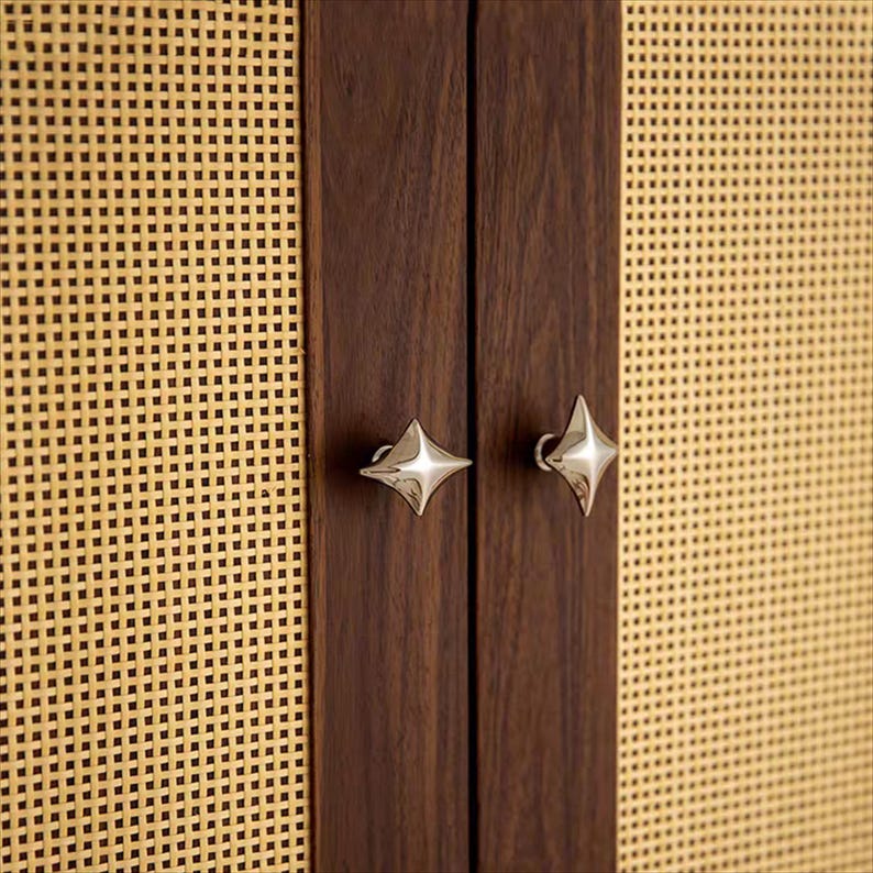 以下が含まれることがあります： 織り込まれた籐のパネルと濃い色の木製のドアを備えたキャビネットドアのクローズアップ。ドアには星型のシルバーの取っ手が付いています。キャビネットはミッドセンチュリーモダンな美学を持っています。