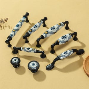 Op de afbeelding: Een verzameling zwarte kastbeslagen, waaronder handgrepen en knoppen. De keramische handgrepen en knoppen hebben een grijs, wit, blauw en geel bloemmotief. De hardware is ontworpen voor gebruik op kasten en laden.