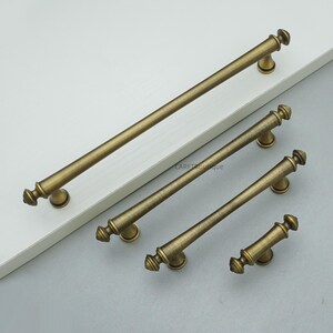 Puede incluir: Cuatro tiradores de armario de latón antiguo de diferentes longitudes están dispuestos sobre una superficie blanca. Cada tirador presenta una barra cilíndrica con tapas decorativas y placas de montaje circulares. Acabado cálido y envejecido.