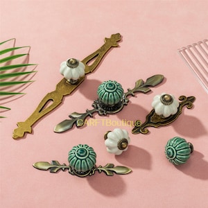 Könnte beinhalten: Eine Auswahl an dekorativer Schrankhardware, darunter Schubladengriffe und Knöpfe. Die Hardware verfügt über Messing- und Antik-Bronze-Oberflächen mit Keramikknöpfen in Weiß und Türkisgrün. Die Knöpfe haben ein Blumendesign.