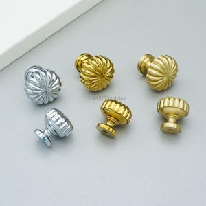 May include: Six decorative cabinet knobs in gold and silver finishes. The knobs have a ribbed, rounded design. The knobs are arranged on a light gray surface. The knobs are likely for furniture or home decor.