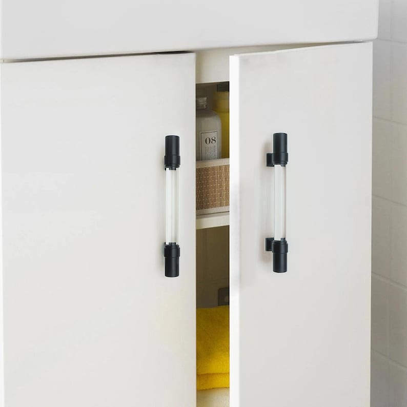 K&ouml;nnte beinhalten: Wei&szlig;e Schrankt&uuml;ren mit schwarzen und transparenten Acryl-Stangengriffen. Die Schrankt&uuml;ren sind leicht ge&ouml;ffnet und geben einen Blick ins Innere frei. Die Griffe sind zylindrisch und vertikal montiert.