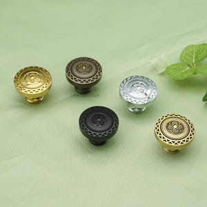 May include: Five decorative cabinet knobs in various metallic finishes: gold, bronze, silver, and black. Each knob features a circular design with a central floral pattern and a geometric border. The knobs are displayed on a light green surface.