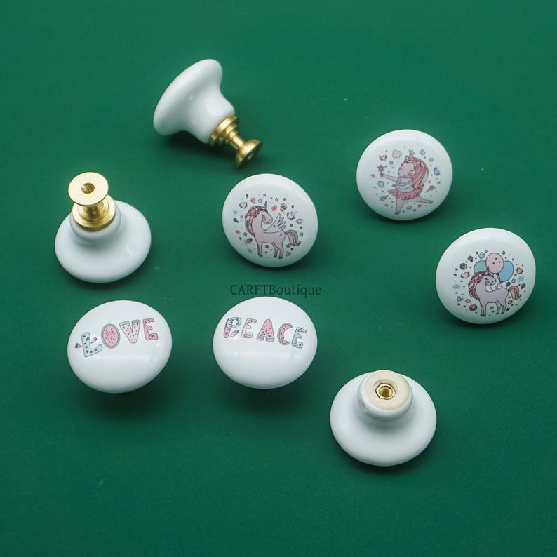 May include: White ceramic cabinet knobs with gold hardware. Some knobs feature unicorn illustrations, while others display the words "LOVE" and "PEACE" in pastel colors. The knobs are set against a green background.
