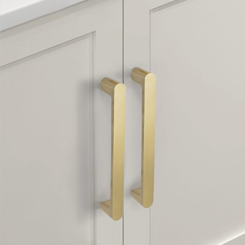 May include: Close-up of two gold-coloured cabinet pulls mounted on a white cabinet door. The pulls are rectangular with rounded edges and a vertical orientation. The cabinet door has a recessed panel design.
