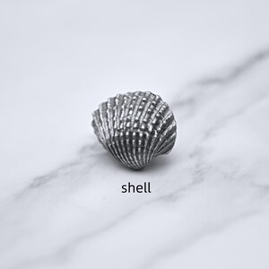Peut inclure: Un coquillage argent&eacute; d&eacute;taill&eacute; avec une texture stri&eacute;e. Le coquillage est pos&eacute; sur une surface de marbre blanc et gris. Le mot "shell" est &eacute;crit en noir, en police sans-serif, en dessous.