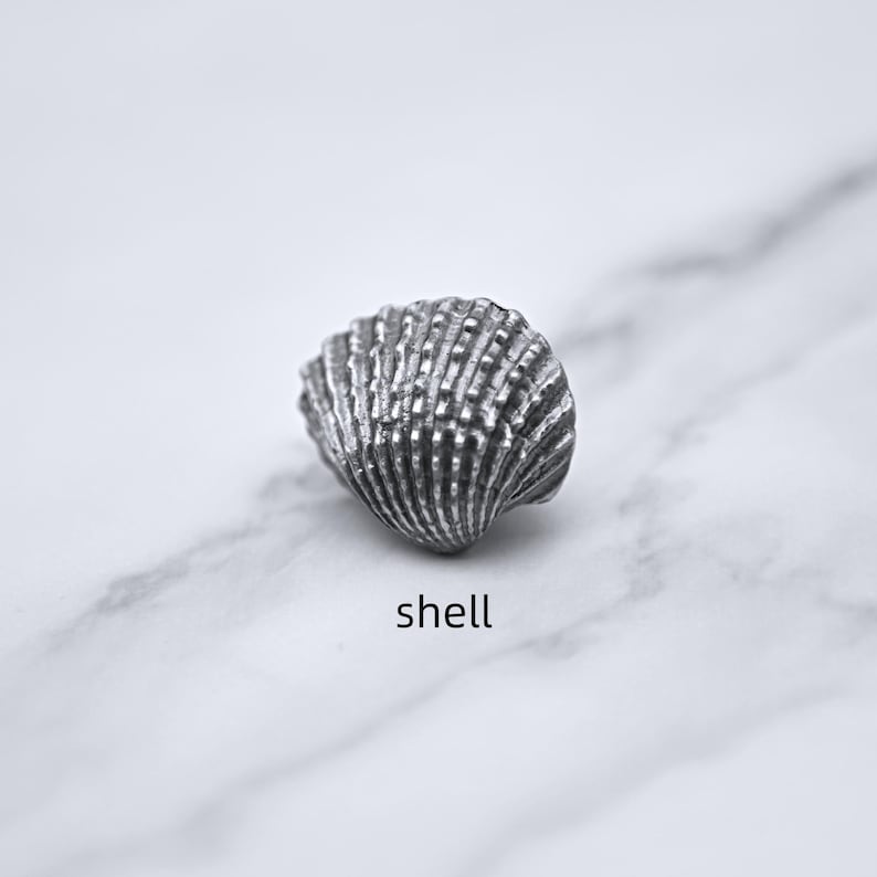Peut inclure: Un coquillage argent&eacute; d&eacute;taill&eacute; avec une texture stri&eacute;e. Le coquillage est pos&eacute; sur une surface de marbre blanc et gris. Le mot "shell" est &eacute;crit en noir, en police sans-serif, en dessous.