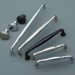 May include: Assortment of cabinet hardware, including silver, black, and brushed nickel handles and knobs. The handles vary in size and design, with curved and straight options. The knobs are round.