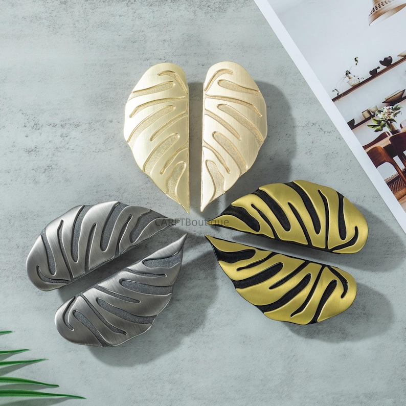 May include: An assortment of decorative leaf-shaped cabinet pulls in various metallic finishes. The pulls are in gold, silver, and a gold and black combination. The leaf design features detailed veining, adding a touch of nature-inspired elegance to furniture.