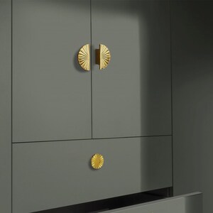 May include: A grey cabinet with gold-coloured hardware. The cabinet doors have half-circle handles, and the drawer has a round knob. The hardware features a sunburst design.