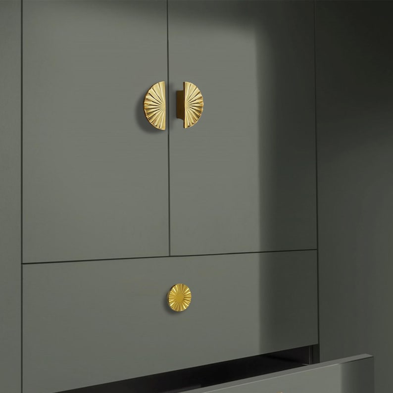 May include: A grey cabinet with gold-coloured hardware. The cabinet doors have half-circle handles, and the drawer has a round knob. The hardware features a sunburst design.