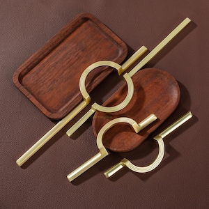 Pode incluir: Uma vista de cima de puxadores de armário dourados e duas bandejas de madeira. Os puxadores têm um design de semicírculo com detalhes lineares e barras retangulares. As bandejas são ovais e retangulares, com acabamento castanho escuro, sobre um fundo castanho.