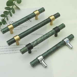 Może przedstawiać: Cztery zielone marmurowe uchwyty do szafek z mosiężnymi, czarnymi i srebrnymi akcentami. Uchwyty są cylindryczne z marmurowym wzorem. Widoczny jest tekst "CARFTBoutique".