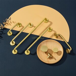 May include: A collection of gold-toned cabinet hardware, including handles and knobs, arranged on a wooden plate and a tan semi-circle against a dark blue background. The hardware has a modern, minimalist design.