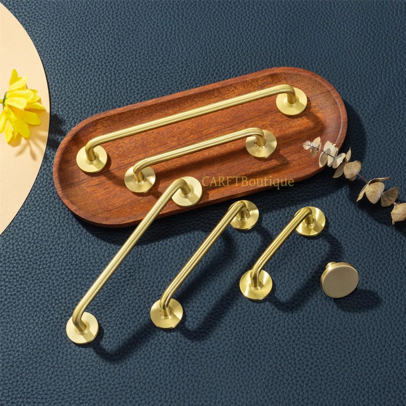 May include: A collection of brass-colored cabinet hardware, including handles and a knob, arranged on a wooden tray. The hardware has a modern, minimalist design with rounded edges and circular mounting plates. The tray sits on a dark blue surface.