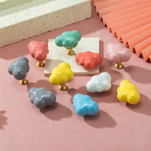 May include: A selection of cloud-shaped cabinet knobs in a range of colours, including pink, green, yellow, blue, and grey. Each knob has a gold-coloured base. The knobs are displayed on a pink surface.