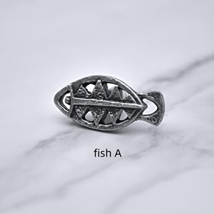 Peut inclure: Une petite &eacute;pingle en m&eacute;tal argent&eacute; en forme de poisson. L'&eacute;pingle pr&eacute;sente un design ajour&eacute; d&eacute;taill&eacute; avec des d&eacute;coupes triangulaires et incurv&eacute;es. Le texte "fish A" est imprim&eacute; en dessous.