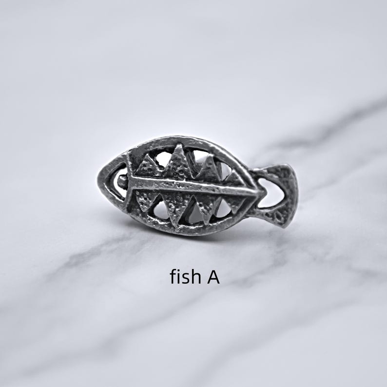 Peut inclure: Une petite &eacute;pingle en m&eacute;tal argent&eacute; en forme de poisson. L'&eacute;pingle pr&eacute;sente un design ajour&eacute; d&eacute;taill&eacute; avec des d&eacute;coupes triangulaires et incurv&eacute;es. Le texte "fish A" est imprim&eacute; en dessous.