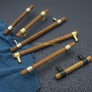 以下が含まれることがあります： 金属アクセントが付いた木製キャビネットプル。プルは、金属リングと金属ポストを備えた円筒形の木製ハンドルを備えています。金属アクセントはシルバー、ゴールド、ブラックです。「CARETBoutique」の文字が見えます。