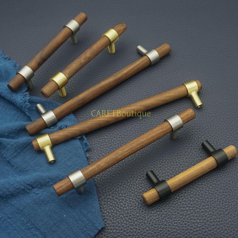 以下が含まれることがあります： 金属アクセントが付いた木製キャビネットプル。プルは、金属リングと金属ポストを備えた円筒形の木製ハンドルを備えています。金属アクセントはシルバー、ゴールド、ブラックです。「CARETBoutique」の文字が見えます。