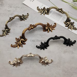 Può includere: Un assortimento di maniglie decorative per mobili in varie finiture. Le maniglie presentano un design curvo e ornato con dettagli a forma di foglia. I colori includono oro, bronzo, nero e argento. Le maniglie sono disposte su una superficie grigio chiaro.
