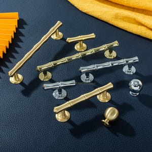 Puede incluir: Una colección de tiradores y pomos dorados y plateados para armarios. Los tiradores tienen un diseño de bambú. La ferretería se muestra sobre una superficie azul oscuro, con una tela amarilla de fondo. La ferretería es de metal.