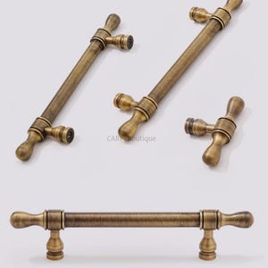 K&ouml;nnte beinhalten: Antike Messing-Schrankgriffe und -kn&ouml;pfe. Die Hardware hat ein T-f&ouml;rmiges Design mit einer zylindrischen Stange und dekorativen Enden. Die Griffe haben ein Vintage-Aussehen mit einer warmen, goldbraunen Farbe. Mehrere St&uuml;cke sind abgebildet.